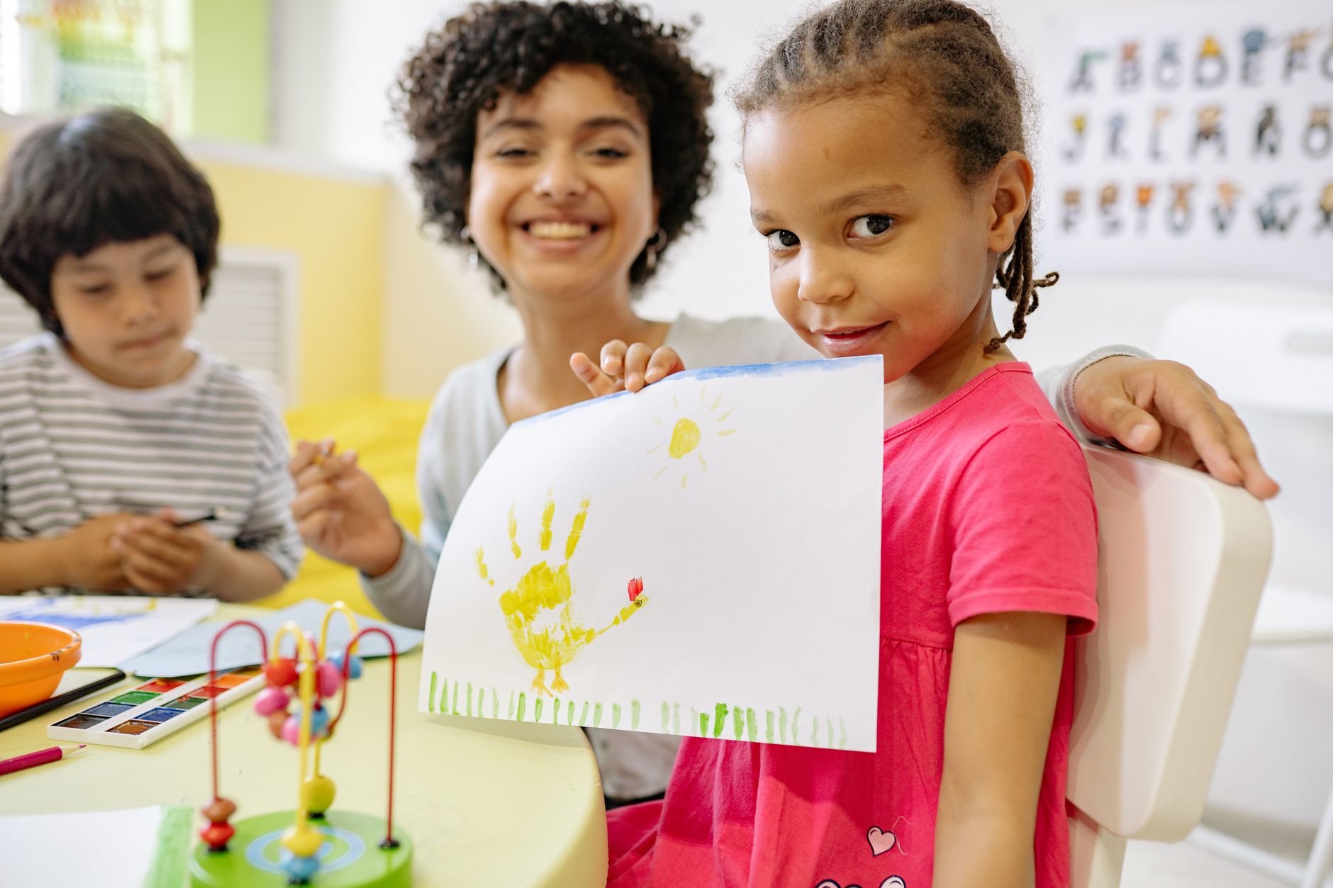 Young children seated at a classroom table, with one child holding up a handprint painting while participating in arts and learning activities.