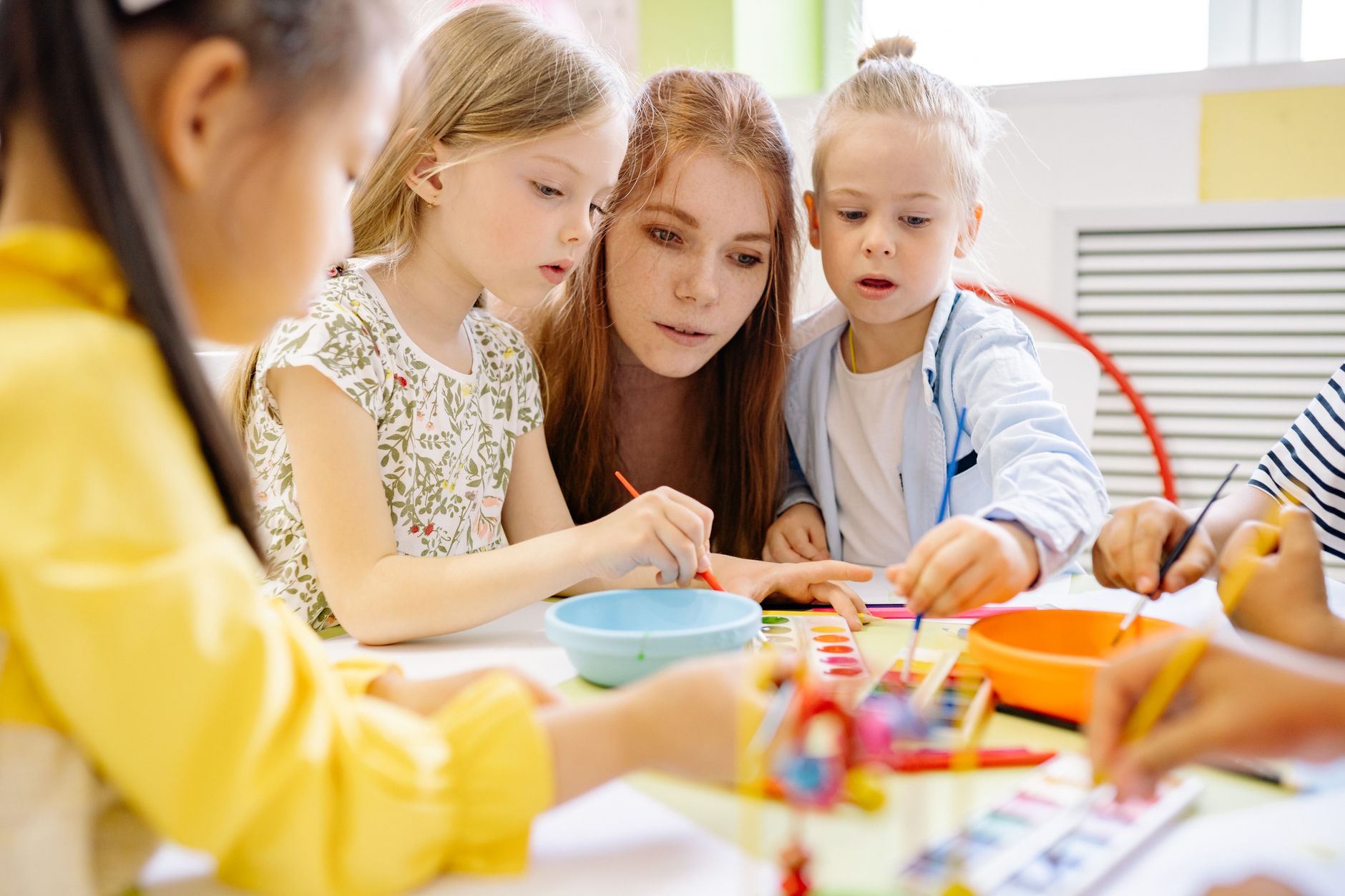 An early childhood educator guiding two young children as they participate in a hands-on painting activity in an early learning classroom.
